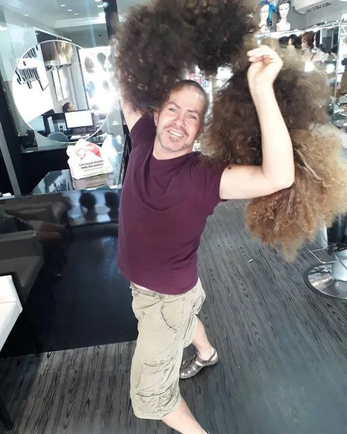 Dirceu Paigel segurando duas perucas de cabelo afro da remessa para a Fundação Laço Rosa, no salão